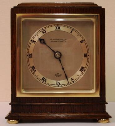 English late c20th Elliott Oak cased 8 day timepiece retailed by J.R.Ogden & Sons Ltd, London & Harrowgate. Stained oak case with square brass surround to matt silvered dial and silvered chapter ring with decorative blued hands, and standing on brass bun feet. The clock displays the standard Elliott back plate with captive, brass hands adjust button and key, and the slow / fast adjuster. Elliott case numbering - 2198 N.