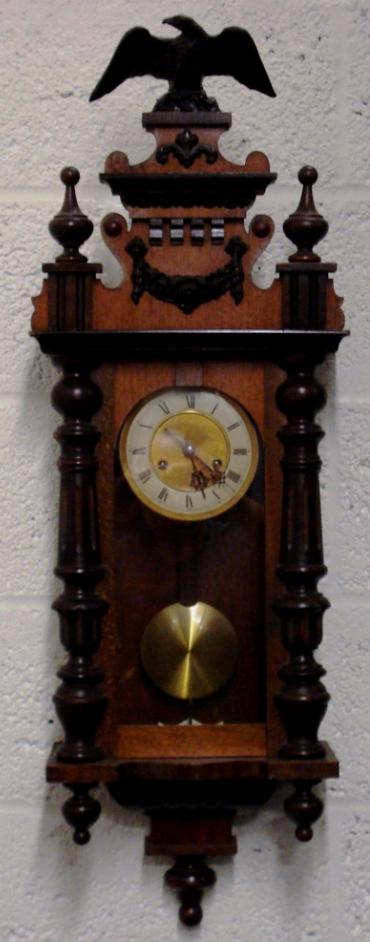 German 8 day gong striking wall clock circa 1900, spring driven pendulum movement housed in an oak and pine case with decorative turned finials and side columns together with applied swags and surmounted by an eagle. Ivory and brass coloured dial with black roman hour markers and black steel hands, and an enamel pendulum regulation plate at the case bottom.