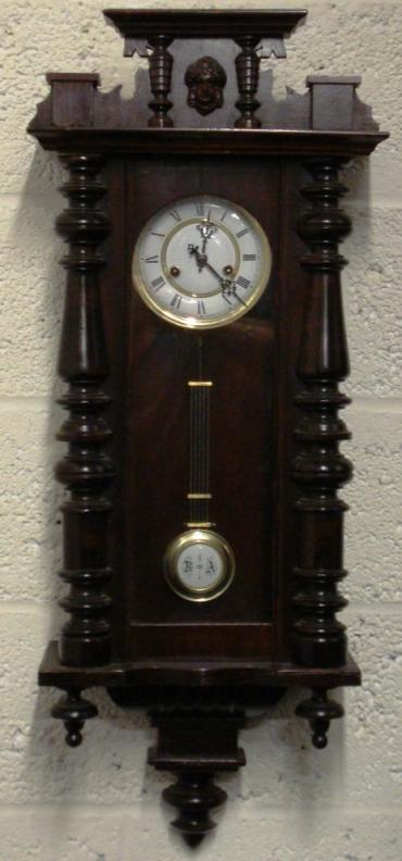 German 8 day gong striking regulator style wall clock circa 1900, with a spring driven pendulum movement housed in a dark pine case with decorative turned finials and side columns together with an applied mask decoration. White enamel and gilded brass dial with black roman hour markers and ornate black steel hands, and a grid iron style pendulum.