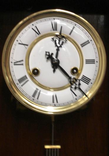 German 8 day gong striking regulator style wall clock circa 1900, with a spring driven pendulum movement housed in a dark pine case with decorative turned finials and side columns together with an applied mask decoration. White enamel and gilded brass dial with black roman hour markers and ornate black steel hands, and a grid iron style pendulum.