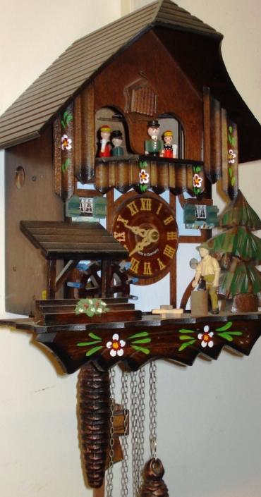 Modern German Automaton musical box day going cuckoo clock. Carved pine wood case with pine tree, water wheel and woodsman decoration. Traditional carved light coloured wood chapter ring with roman hours and matching wooden hands. Visible pendulum and triple cast metal pine cone weights. On the hour the carved wood cuckoo bird displays the hours, and the mechanism activates the figure carousel, the woodman chopping and the water wheel turning.