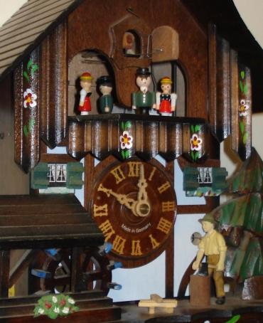Modern German Automaton musical box day going cuckoo clock. Carved pine wood case with pine tree, water wheel and woodsman decoration. Traditional carved light coloured wood chapter ring with roman hours and matching wooden hands. Visible pendulum and triple cast metal pine cone weights. On the hour the carved wood cuckoo bird displays the hours, and the mechanism activates the figure carousel, the woodman chopping and the water wheel turning.