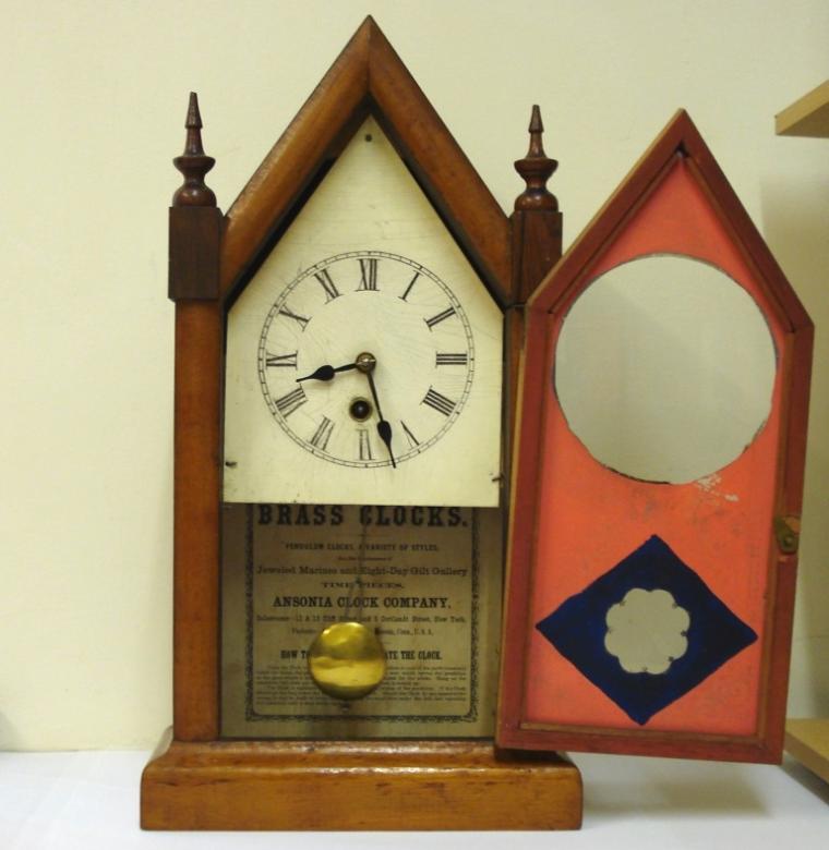 American 30 hour fruit wood veneer cased mantel clock timepiece circa 1885. Simple lancet topped case with turned finials and integral plinth. Full length lancet topped door with flat glass painted with pink and blue decoration and over an aged white painted dial plate with black roman hours and black steel hands. Simple american Ansonia Clock Co. brass, spring driven pendulum regulated, movement with moving pendulum visible through the glass door.