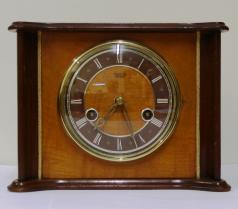 English Smiths Enfield 8 day wood veneer cased gong strike mantel clock circa 1950. Unusually shaped flat top case with gilt bezel and domed glass over a dark brown chapter ring with white roman hours and pierced gilt hands. Rear door to brass spring driven, floating balance movement with slow / fast regulation. Time adjust and winding squares behind the front face glass. Dimensions: Height - 6.75", width - 9", depth - 5".