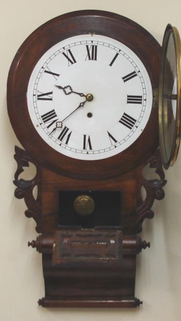 American Ansonia Clock Co. round faced extended drop dial 8 day wall clock in a carved and moulded mahogany case with scroll bottom. Brass bezel with flat glass over a white painted dial with black roman numerals and ornate black painted hands with separate lower in view brass pendulum window. Spring driven, pendulum regulated, time piece movement, circa 1890.  Dimensions: Height - 31", Width - 16.5", Depth 4.5".