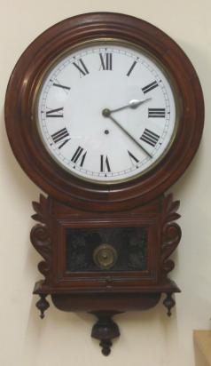 English 8 day wall clock by the British United Clock Co. Ltd of Birmingham. Round mahogany case with brass bezel and flat glass over a white painted dial with black Roman hours and polished silvered hands. Lower extended drop dial with carved and moulded casework and visible pendulum window. Signed basic brass spring driven, pendulum regulated, time piece movement, circa 1900.  Dimensions: Height - 30", Width - 10.5", Depth 5".