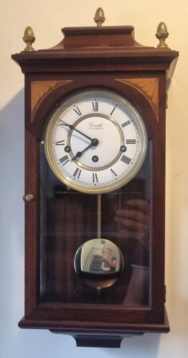 Modern 8 day Westminster chime wall clock by Comitti of London. Mahogany caddy top case with decorative inlay and gilt brass acorn finials. Flat glass front and sides with a key wound, spring driven, brass pendulum regulated movement. White dial with gilt bezel and black Roman hours with black painted hands. Dimensions: Height - 20", Width - 8.5", Depth - 5".
