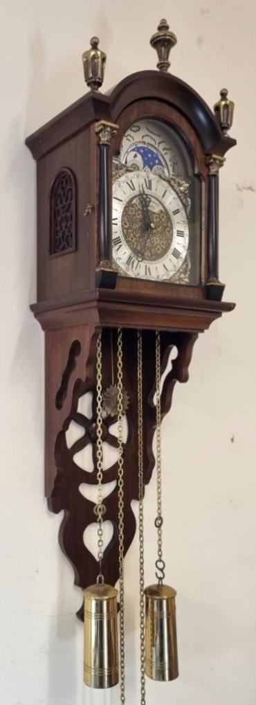Modern Reproduction Dutch style wall clock by St James of London. Mahogany hooded top case with brass finials and opening front with brass mounted side columns and fret worked side panels and lower bracket base. Flat glass front over highly decorated brass dial plate with silvered chapter ring and black Roman hours and hands together with applied angel spandrels all surmounted by a lunar calendar and moon phase indicating panel. German 8 day weight driven, pendulum regulated movement striking the half hour and hour on a bell, with decorative sun burst pendulum and turned polished brass weights. Case Dimensions: Height - 27", Width - 8", Depth - 6".