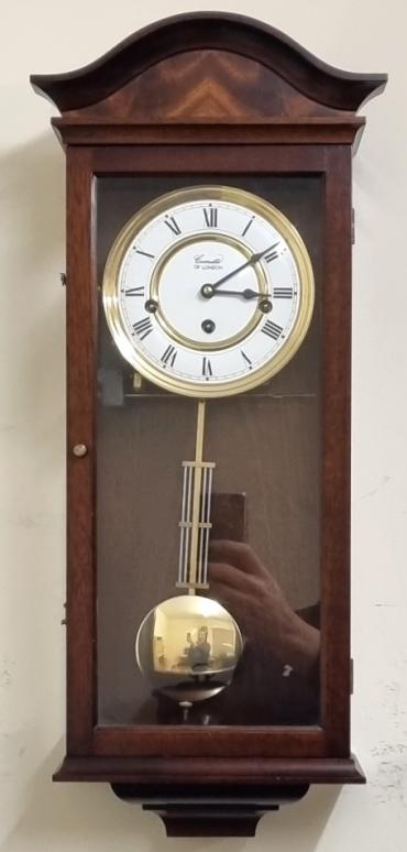Modern 8 day Westminster chime wall clock by Comitti of London. Mahogany case with decorative veneered scroll top and opening glass front with two glass side panels. Round brass bezel and white dial plate with black Roman hours and matching hands together with lower polished brass pendulum. 8 day spring driven, pendulum regulated movement striking the quarters and the hour on horizontally mounted steel rods.  Case Dimensions: Height - 22", Width - 9", Depth - 5".