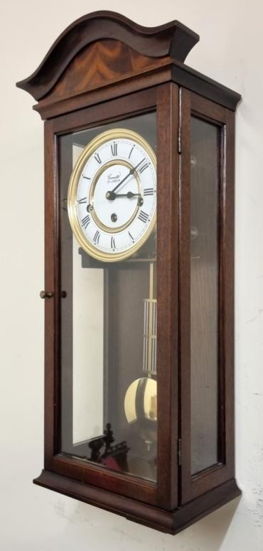 Modern 8 day Westminster chime wall clock by Comitti of London. Mahogany case with decorative veneered scroll top and opening glass front with two glass side panels. Round brass bezel and white dial plate with black Roman hours and matching hands together with lower polished brass pendulum. 8 day spring driven, pendulum regulated movement striking the quarters and the hour on horizontally mounted steel rods.  Case Dimensions: Height - 22", Width - 9", Depth - 5".